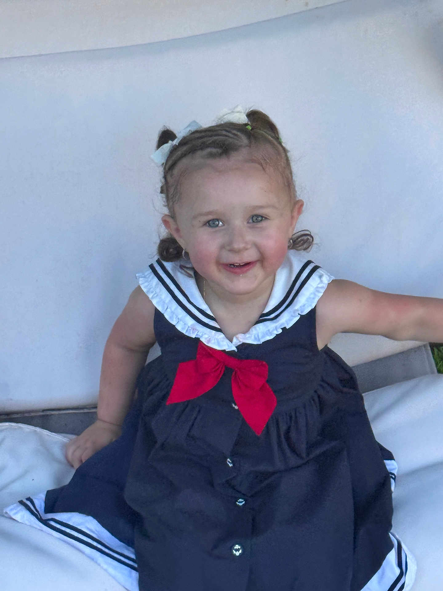 Laylani is registered to the contest to win money with this photo: child, toddler, smiling, dress, sailor_style, bow, blue_eyes, curly_hair, hair_bows, sitting, white_cushion, outdoor, happy, cute, portrait, person, young_child, fashion, buttoned_dress, skin