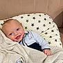 baby, smiling, blue_eyes, blanket, knitted_blanket, cushion, star_pattern, infant, cute, comfort, indoors, soft_furniture, child, portrait, happy, cozy, person, head, clothing, pale_pink_background