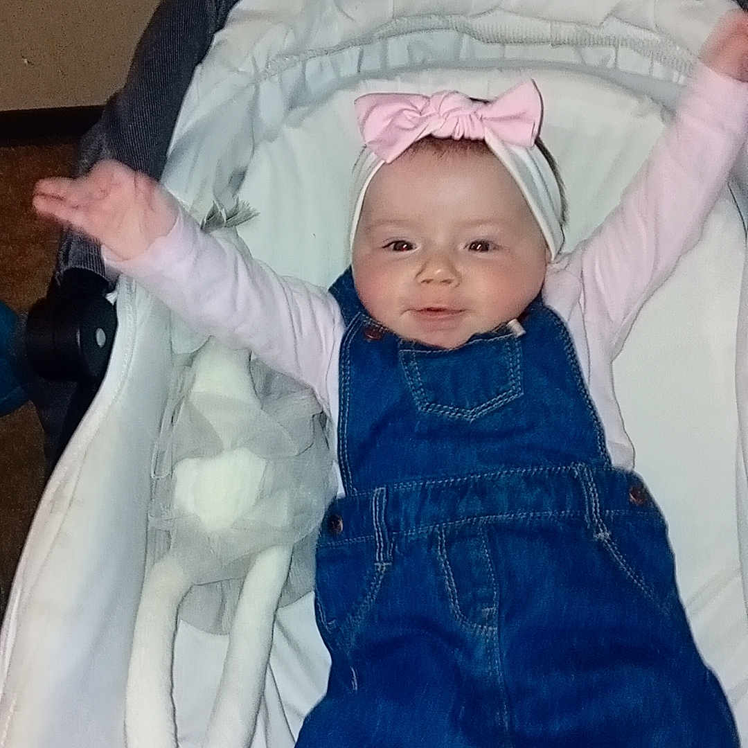 Lya participe au concours pour gagner de l'argent avec cette photo : baby, baby_seat, blanket, child, clothing, cozy, cute, denim_dress, face, happy, headband, indoor, infant, person, pink_bow, raising_hands, smiling, soft_toys, toddler, toy