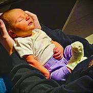Mckenna joined the competition — help win amazing prizes! baby, infant, newborn, sleeping, cradled, adult_hands, onesie, white_shirt, purple_pants, sock, cozy, portrait, indoor, warm_light, lap, black_sweatshirt, peaceful, skin, floor_tiles, handheld