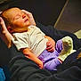 baby, infant, newborn, sleeping, cradled, adult_hands, onesie, white_shirt, purple_pants, sock, cozy, portrait, indoor, warm_light, lap, black_sweatshirt, peaceful, skin, floor_tiles, handheld