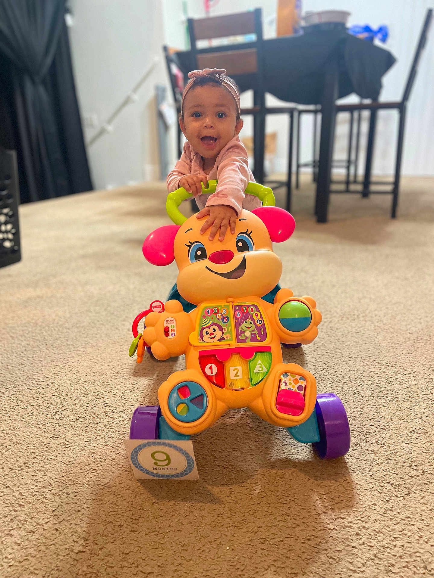Olivia is registered to the contest to win money with this photo: baby, child, toy, walker, carpet, indoor, pink_headband, smile, happy, colorful, furniture, table, chair, floor, playtime, infant, person, cute, expression, young