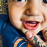 Ace joined the competition — help win amazing prizes! baby, close_up, smiling, teeth, blue_shirt, tattoo, hand, face, child, person, cute, young, eyes, skin, mouth, expression, indoor, human, portrait, infant