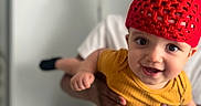 Ace joined the competition — help win amazing prizes! baby, child, red_hat, yellow_shirt, smiling, indoor, person, hand, face, adult, knitted_hat, happy, cute, holding, portrait, playful, clothing, expression, background, focus