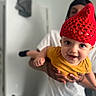 baby, child, red_hat, yellow_shirt, smiling, indoor, person, hand, face, adult, knitted_hat, happy, cute, holding, portrait, playful, clothing, expression, background, focus