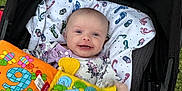 Harlow is registered to the contest to win money with this photo: baby, smiling, stroller, outdoor, soft_book, toy, grass, blanket, child, infant, colorful, happy, person, face, clothing, play, seat, comfort, cute, young