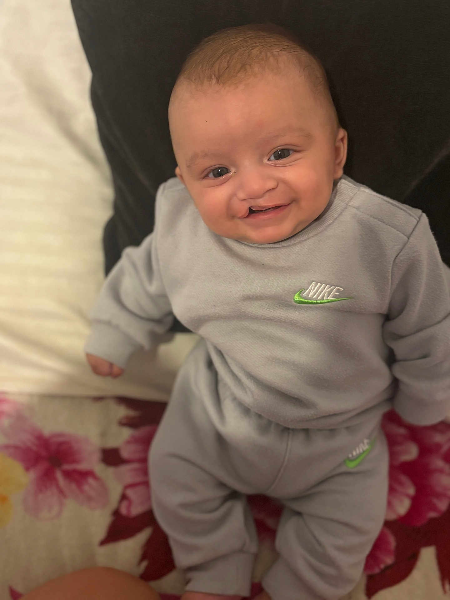 Denis is registered to the contest to win money with this photo: baby, smiling, infant, clothing, nike, gray_outfit, sitting, pillow, blanket, floral_pattern, indoor, cute, happy, child, portrait, apparel, young, person, comfort, cozy