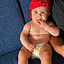 baby, child, red_cap, diaper, blue_couch, sitting, fingers_in_mouth, cute, curious, skin, arm, indoor, person, infant, casual, relaxed, young, portrait, headwear, comfort