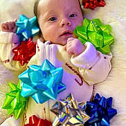Kaia joined the competition — help win amazing prizes! baby, infant, face, person, gift_bows, colorful, decorations, holiday, cute, soft_texture, fluffy, white_background, clothing, lying_down, closeup, small_hands, expression, indoors, festive, cozy