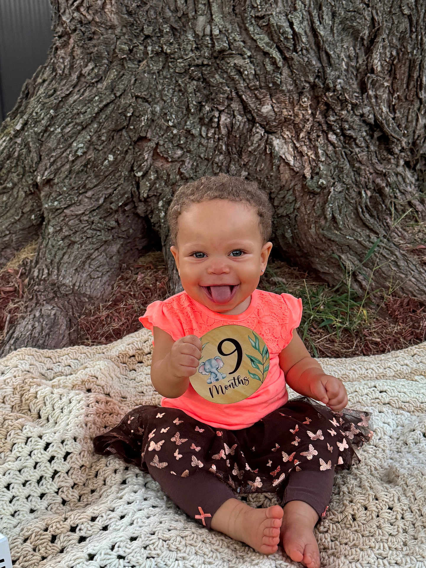 Samarah is registered to the contest to win money with this photo: baby, blanket, child, clothing, cute, face, grass, happy, infant, leggings, nature, outdoor, person, playful, portrait, sitting, skirt, smiling, tongue_out, tree_trunk