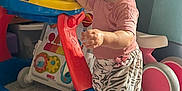 Samarah is registered to the contest to win money with this photo: barefoot, carpet, child, colorful, curly_hair, furniture, happy, indoor, pants, person, pink_shirt, plastic, play, smiling, storage, toddler, toy, walker, wheel, zebra_print