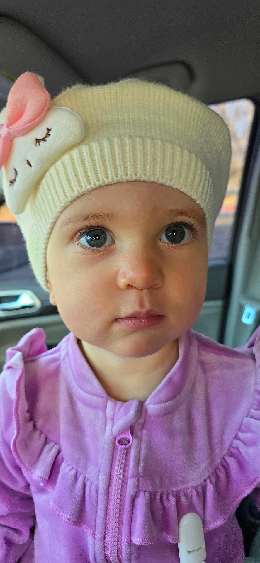 Lia is registered to the contest to win money with this photo: toddler, child, face, hat, knit_hat, pink_bow, purple_jacket, clothing, closeup, indoors, car_interior, curious, innocent_expression, young_child, portrait, winter_clothing, cute, baby, person, eyelashes