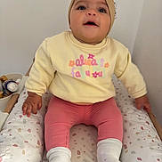 Juna a rejoint le concours — aidez-le/la à gagner de superbes lots ! baby, child, infant, sitting, headband, yellow_sweatshirt, pink_pants, white_socks, cushion, patterned_cushion, indoor, cute, portrait, person, clothing, face, smiling, hands, feet, happy