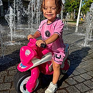 Gianni a rejoint le concours — aidez-le/la à gagner de superbes lots ! child, curly_hair, daylight, fun, happy, outdoor, pavement, person, pink_clothing, play, recreation, smiling, sneakers, summer, sunny, toddler, toy_motorcycle, urban, water_fountain, water_splash