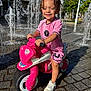 child, curly_hair, daylight, fun, happy, outdoor, pavement, person, pink_clothing, play, recreation, smiling, sneakers, summer, sunny, toddler, toy_motorcycle, urban, water_fountain, water_splash
