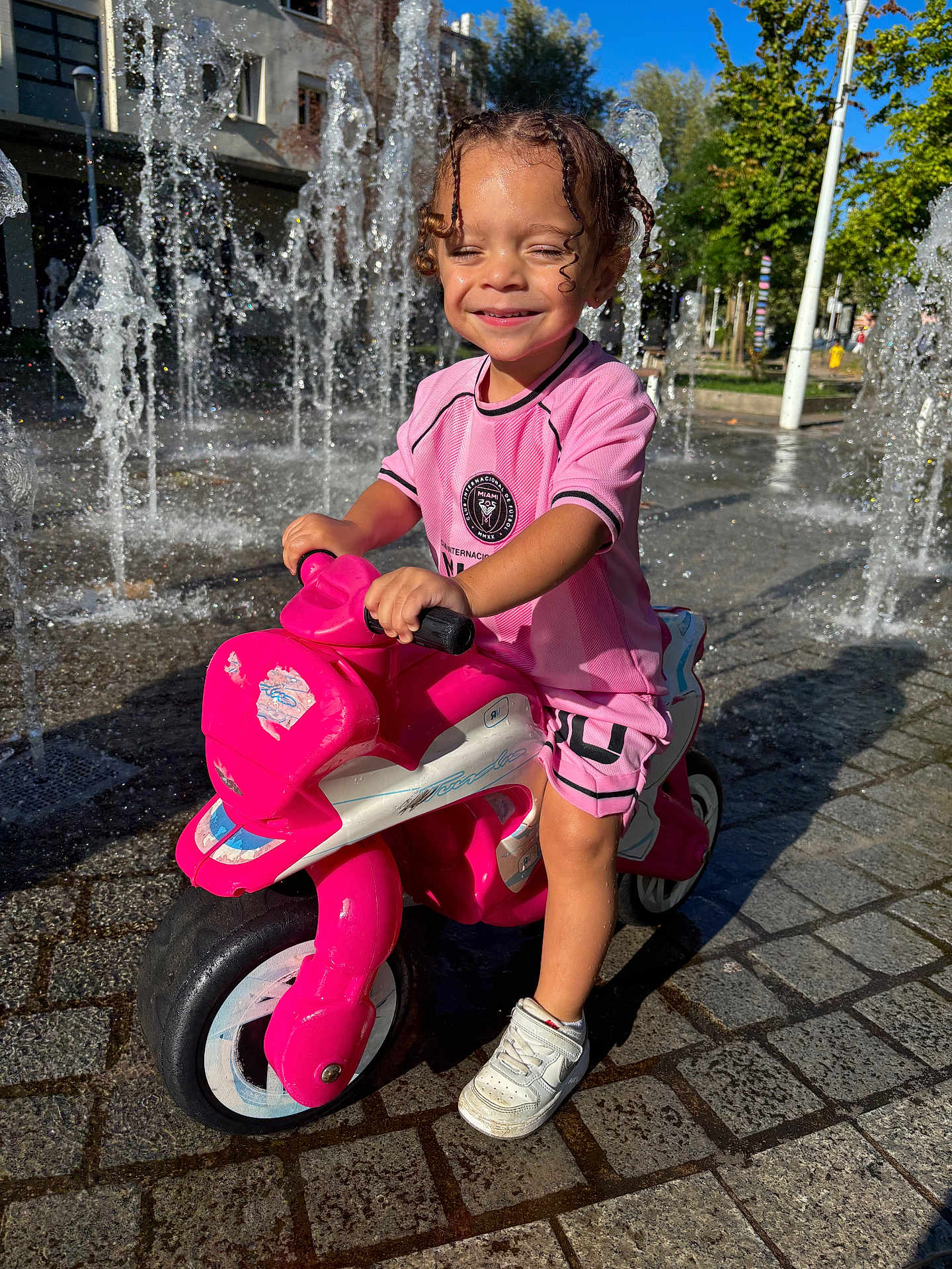 Gianni a rejoint le concours — aidez-le/la à gagner de superbes lots ! child, curly_hair, daylight, fun, happy, outdoor, pavement, person, pink_clothing, play, recreation, smiling, sneakers, summer, sunny, toddler, toy_motorcycle, urban, water_fountain, water_splash