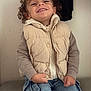 beige_vest, blue_jeans, candid, casual_clothing, child, curly_hair, cute, face, fashion, hands, happy, indoor, joyful, person, portrait, sitting, smiling, toddler, warm_clothing, young_child