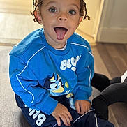 Gianni participe au concours pour gagner de l'argent avec cette photo : blue_sweatshirt, cartoon_character, casual, child, clothing, cute, expression, floor, hair, happy, indoor, person, playful, portrait, sitting, smiling, sneakers, toddler, tongue_out, young