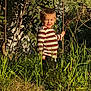 child, smiling, striped_shirt, long_sleeve, pants, grass, plants, trees, outdoor, sunlight, nature, greenery, holding_branch, standing, young, happy, face, daylight, bushes, casual_clothing