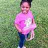 child, girl, smiling, outdoor, grass, greenery, pink_shirt, jeans, sneakers, purse, hair_buns, happy, cute, person, standing, nature, daylight, casual_clothing, young_child, playful