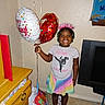 child, birthday, balloons, smile, tiara, tshirt, rainbow_skirt, gift_bag, indoor, wooden_dresser, television, barefoot, happy, girl, celebration, party, hair_beads, floor_tiles, wall, present