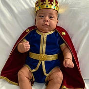 Elian Mateo Leiva A. joined the competition — help win amazing prizes! baby, child, crown, cape, royal, costume, blue_clothing, gold_trim, sitting, indoor, white_couch, cute, portrait, infant, furniture, person, expression, adorable, chubby, hands