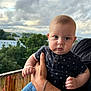 baby, child, person, face, hand, clothing, bracelet, blue_jeans, outdoor, balcony, sky, clouds, trees, nature, portrait, expression, anchor_pattern, holding, adult_hand, casual