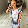 child, smiling, peace_sign, striped_dress, barefoot, indoor, tiled_floor, dining_table, chairs, bicycle, framed_photos, lamp, bracelet, happy, person, portrait, home, wooden_furniture, light, floor