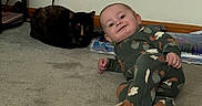 Grayson is registered to the contest to win money with this photo: baby, infant, child, smiling, sitting, onesie, animal_print, cat, tortoiseshell_cat, pet, carpet, indoor, home, living_room, wall, electrical_outlet, books, floor, portrait, adorable