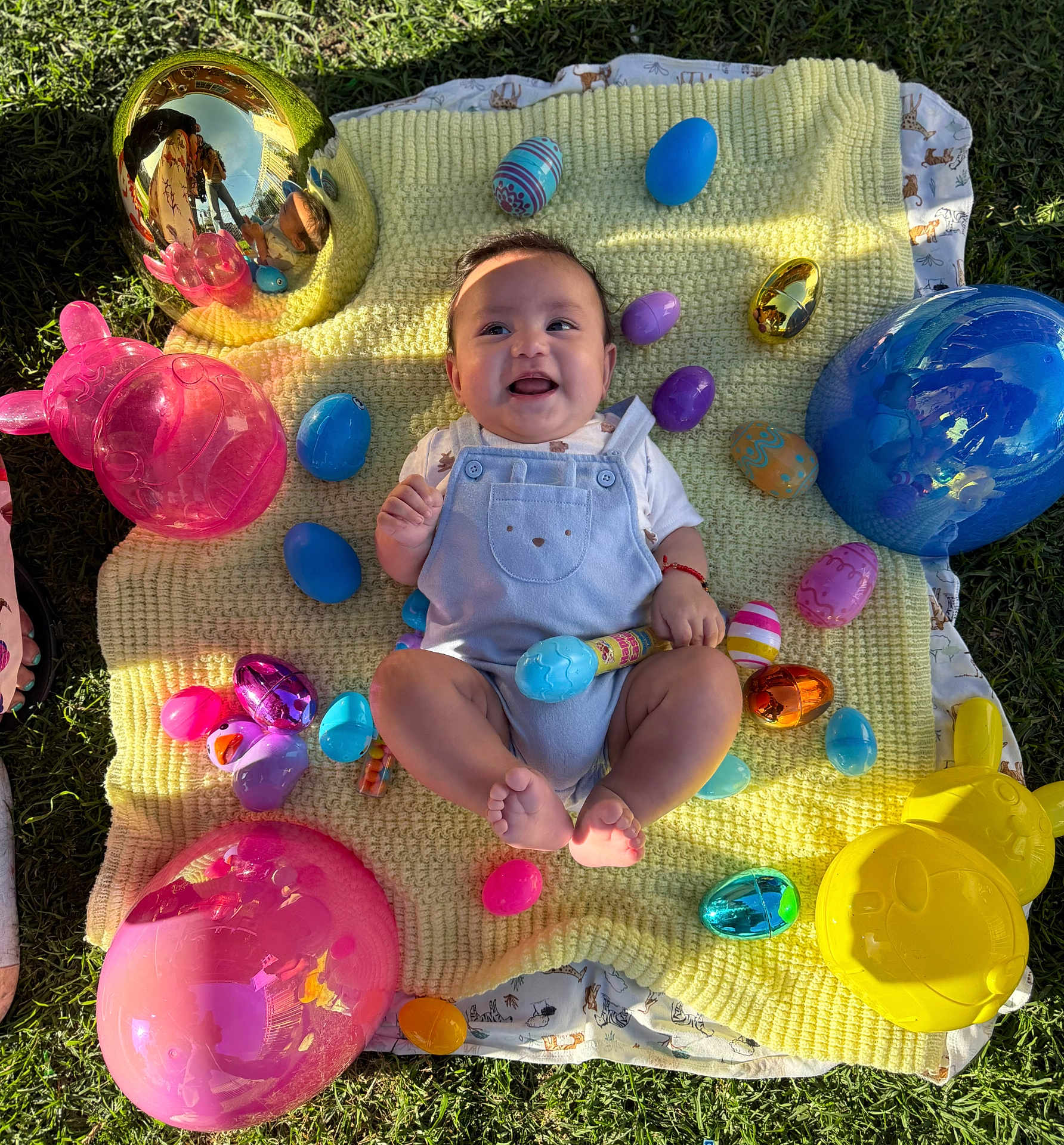 Marcos is registered to the contest to win money with this photo: baby, child, smiling, easter_eggs, balloons, yellow_blanket, grass, outdoor, toy, blue_onesie, colorful, sunlight, happy, sitting, playful, cute, celebration, spring, holiday, festive