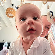 Liam a rejoint le concours — aidez-le/la à gagner de superbes lots ! baby, adult, face, close_up, indoor, toy, mobile, smile, curious, person, child, white_clothing, window, sunlight, home, hanging_toys, playful, head, expression, background