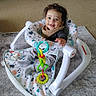 baby, child, smiling, baby_walker, toy, indoor, carpet, bib, clothing, happy, face, curly_hair, seat, playtime, infant, person, floor, home, baby_toy, young_child