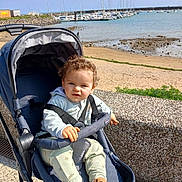 Maël a rejoint le concours — aidez-le/la à gagner de superbes lots ! toddler, stroller, child, outdoor, beach, water, boats, sky, sunny, casual_clothing, curly_hair, footwear, seaside, harbor, concrete_wall, greenery, daylight, person, safety_strap, marine