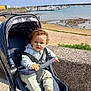 toddler, stroller, child, outdoor, beach, water, boats, sky, sunny, casual_clothing, curly_hair, footwear, seaside, harbor, concrete_wall, greenery, daylight, person, safety_strap, marine