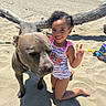 child, dog, beach, ocean, sand, driftwood, outdoor, smiling, peace_sign, swimsuit, curly_hair, kneeling, pet, pit_bull, animal, summer, sunlight, shadows, beach_toys, portrait