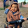 baby, blue_shirt, brown_clothing, casual_clothing, child, cute, daylight, fun, handlebar, happy, hooded_poncho, outdoor, play, portrait, smiling, toddler, toy, transport, tricycle, young_child