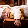 baby, sleeping, white_outfit, knit_hat, wooden_buttons, teddy_bear, brown_cushion, soft_texture, indoor, cozy, child, peaceful, cute, resting, portrait, hand, toy, warm_lighting, close_up, infant