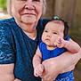 elderly_woman, baby, blue_clothing, outdoor, portrait, holding, family, love, affection, person, skin, head, face, nature, tree, casual_wear, smile, tenderness, closeup, human