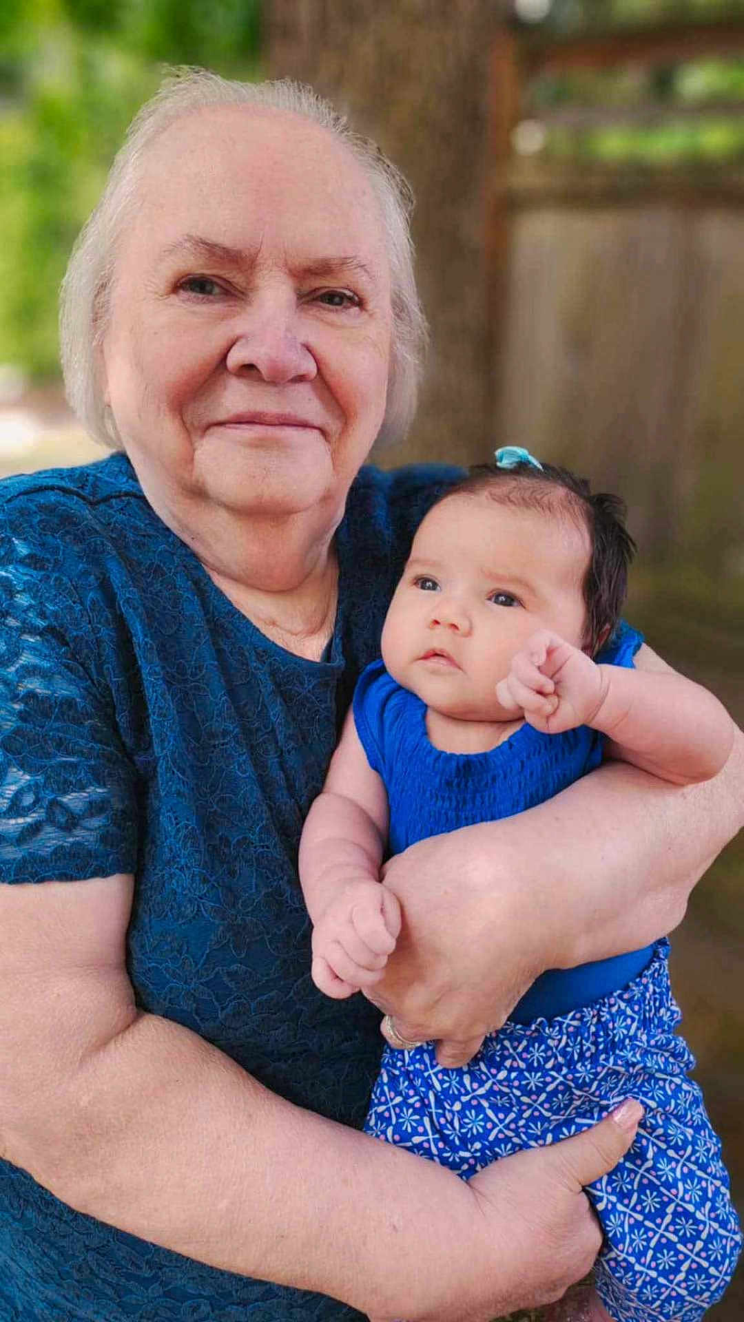 Raziah Morgan is registered to the contest to win money with this photo: elderly_woman, baby, blue_clothing, outdoor, portrait, holding, family, love, affection, person, skin, head, face, nature, tree, casual_wear, smile, tenderness, closeup, human