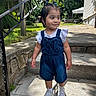 child, toddler, girl, portrait, outdoor, standing, steps, house, garden, sunlight, denim_overalls, ruffled_shirt, sneakers, hair_bows, dark_hair, smiling, side_glance, residential, railing, daytime