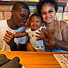Amoni is registered to the contest to win money with this photo: family, baby, smile, happy, mother, father, tattoo, restaurant, wooden_table, curly_hair, casual_clothing, indoor, portrait, people, table, menu, napkin, holding, affection, joy