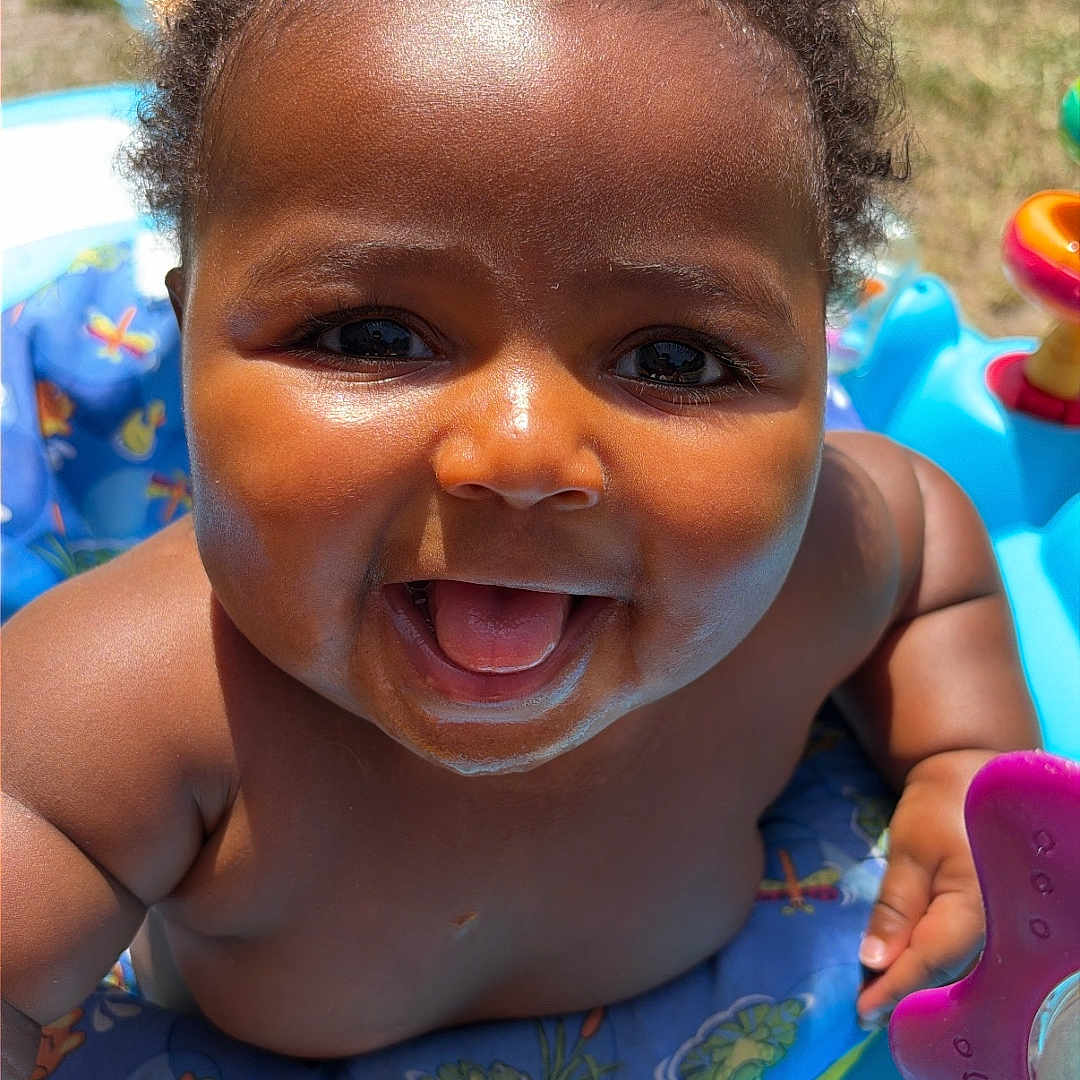 Amoni is registered to the contest to win money with this photo: baby, smiling, outdoor, sunlight, activity_center, child, happy, face, skin, curly_hair, playtime, toy, colorful, summer, infant, closeup, bright, joy, young_child, fun