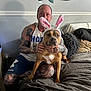 animal, bed, blanket, brown_dog, bunny_ears, casual, dog, friendship, furniture, headband, human, indoor, man, mirror, pet, pillow, relaxed, smile, tattoos, wall