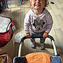 box, casual_clothing, chair, child, curly_hair, expression, floor, furniture, happy, hoodie, indoor, person, plastic_bag, playroom, smiling, sneakers, stroller, toddler, toy, wooden_floor