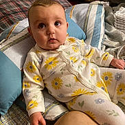Harper is registered to the contest to win money with this photo: baby, infant, onesie, floral_pattern, bed, pillow, blanket, indoor, child, person, cute, sleepwear, soft_lighting, curious, young_child, portrait, cozy, relaxed, hand, face