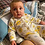 baby, infant, onesie, floral_pattern, bed, pillow, blanket, indoor, child, person, cute, sleepwear, soft_lighting, curious, young_child, portrait, cozy, relaxed, hand, face