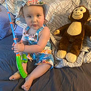 Oliver is registered to the contest to win money with this photo: toddler, child, toy, fishing_rod, stuffed_animal, monkey_toy, bed, blanket, clothing, hat, indoor, person, cute, sitting, playful, colorful, soft_toy, happy, young_child, comfort
