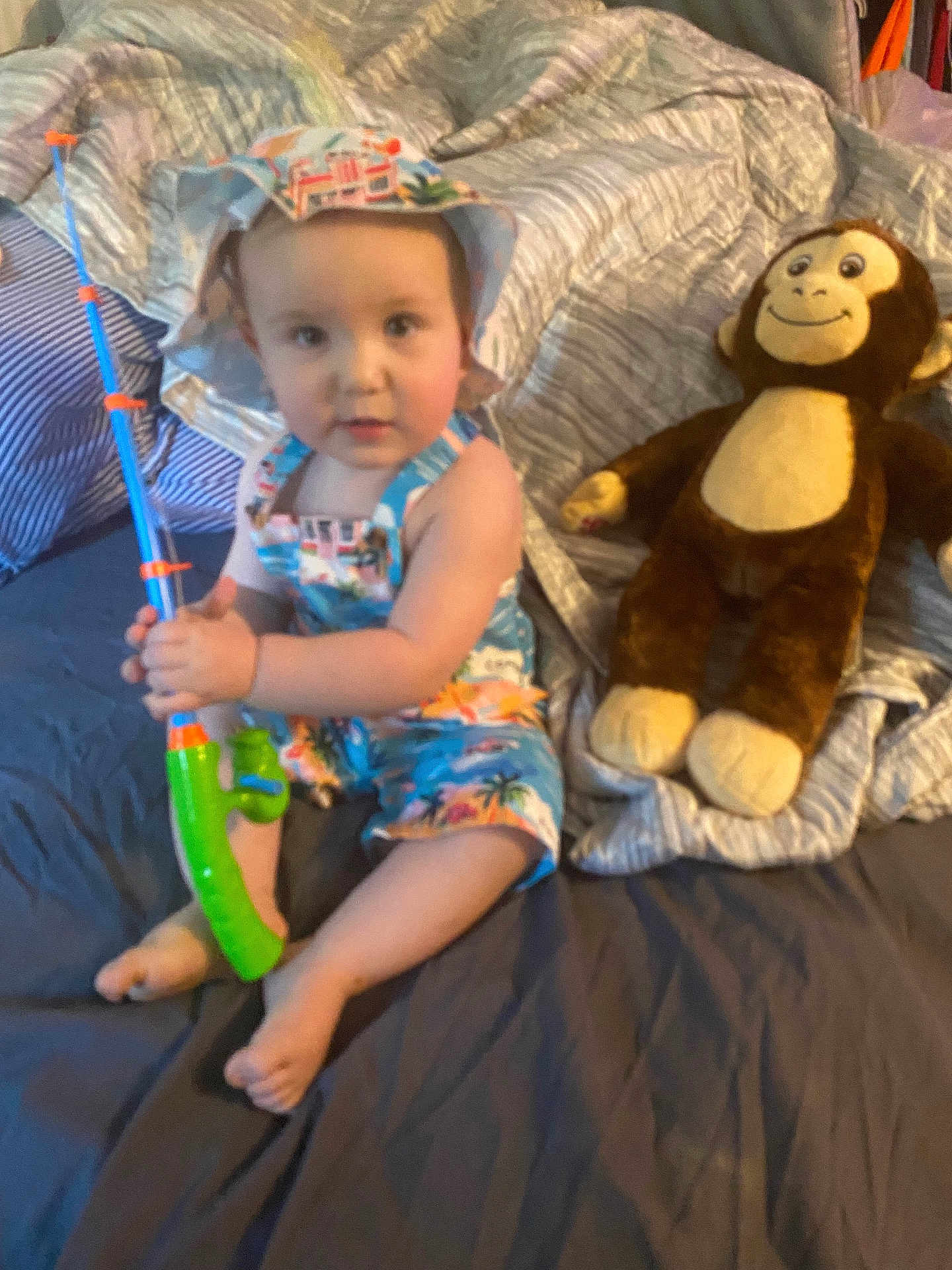 Oliver is registered to the contest to win money with this photo: toddler, child, toy, fishing_rod, stuffed_animal, monkey_toy, bed, blanket, clothing, hat, indoor, person, cute, sitting, playful, colorful, soft_toy, happy, young_child, comfort