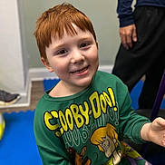 Declan is registered to the contest to win money with this photo: child, red_hair, smile, green_sweater, scooby_doo, indoor, person, seated, blue_mat, adult, hand, casual_clothing, flooring, wall, head, face, long_sleeve, happy, young, portrait