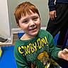 child, red_hair, smile, green_sweater, scooby_doo, indoor, person, seated, blue_mat, adult, hand, casual_clothing, flooring, wall, head, face, long_sleeve, happy, young, portrait